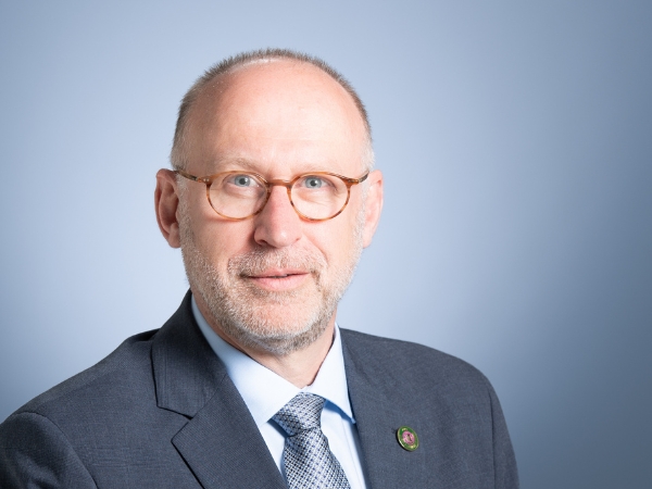 A headshot of a white older man wearing glasses and a grey suit in front of a blue background